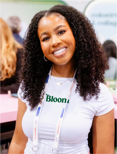 A smiling person with curly hair wearing a white shirt with "Bloom" printed on it, standing in a vibrant event setting.