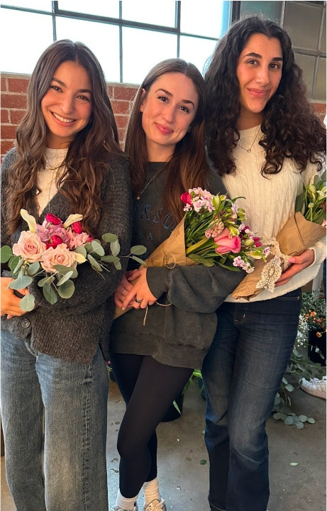 Three women stand together, each holding a bouquet of flowers, smiling in a bright, floral setting.