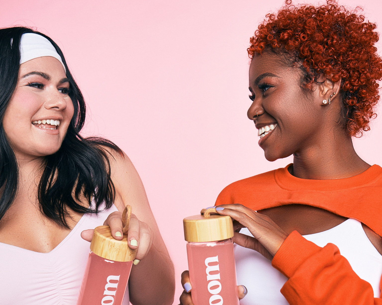 Two smiling women holding pink Bloom drinks in bamboo-lid bottles against a light pink background